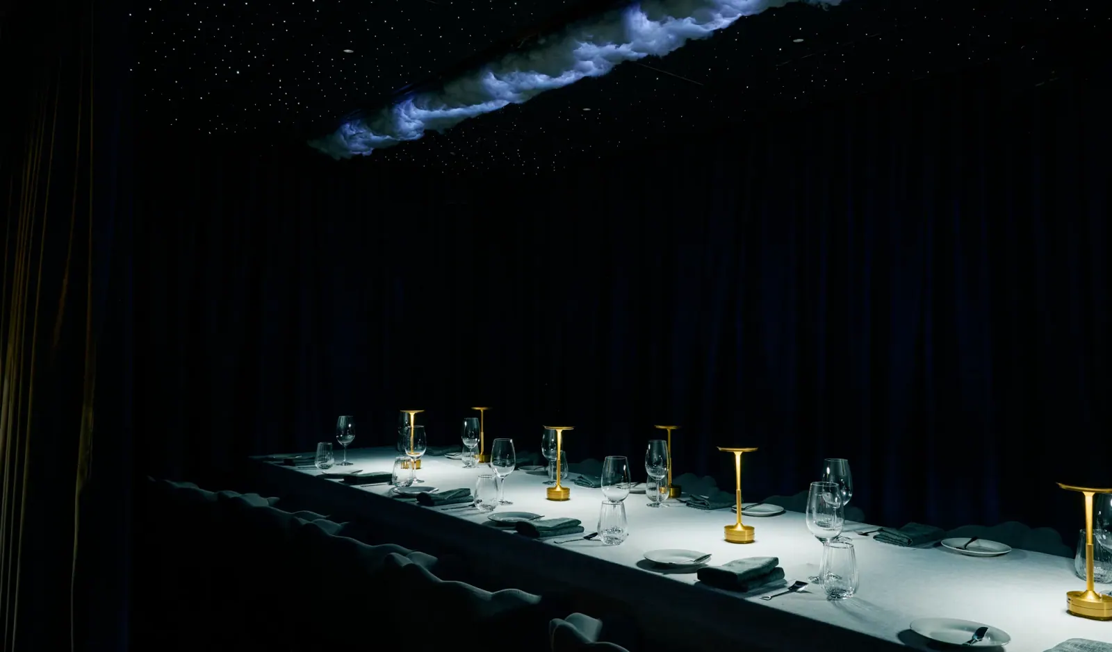 The long table set in the dark, lit from above beneath a drifting cloud